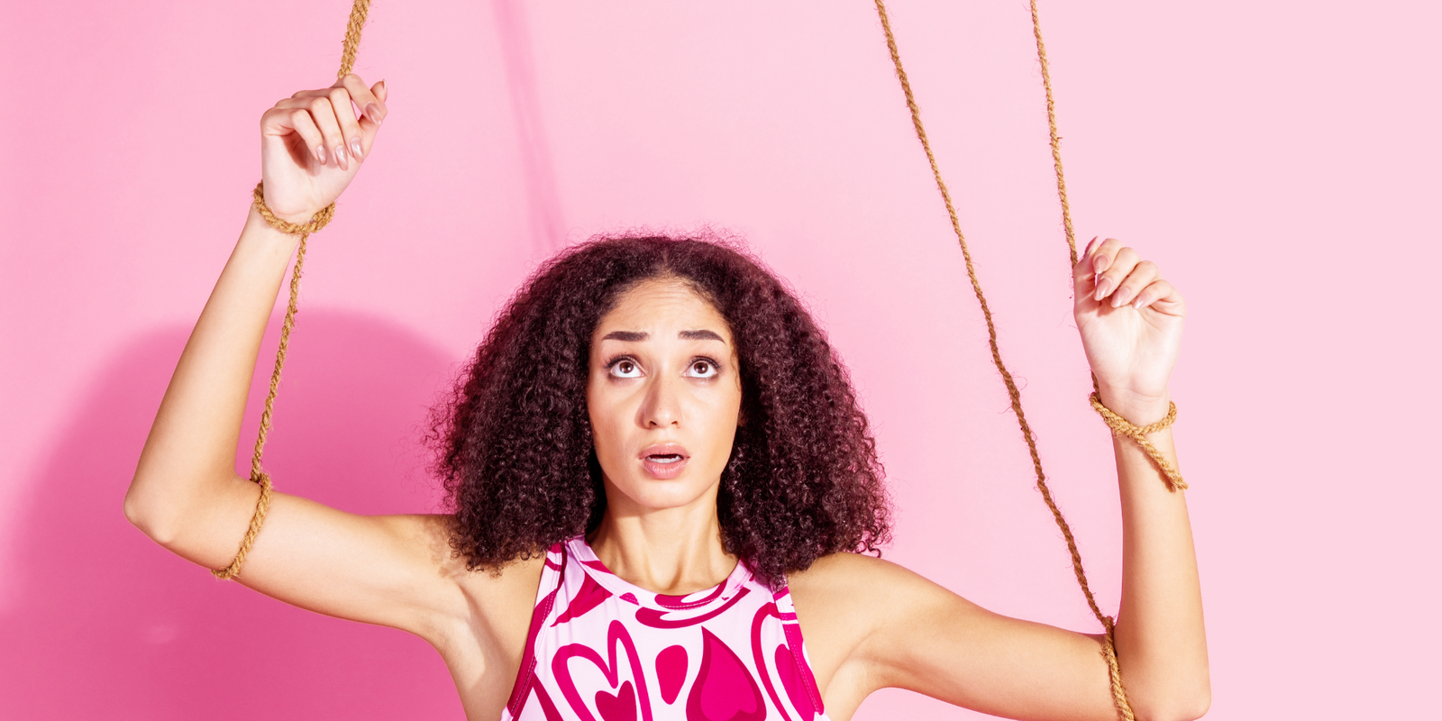 Young woman in a pink patterned top looking upward while holding ropes tied to her wrists like puppet strings, symbolizing the loss of autonomy under beauty and body standards.